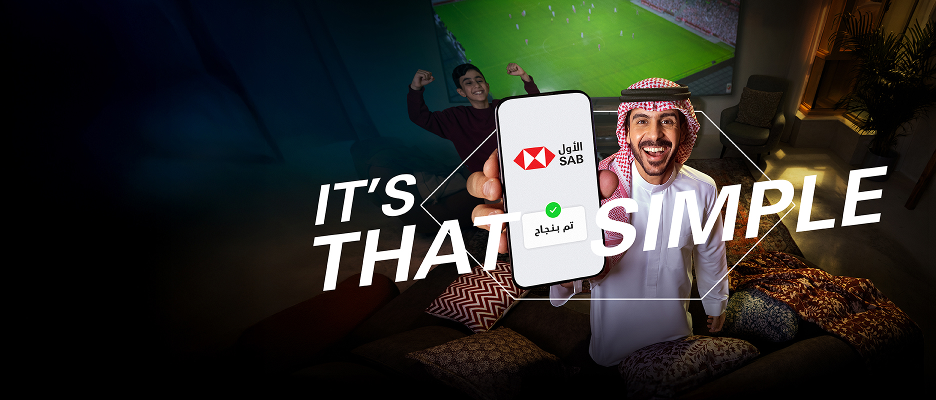Saudi guy in a living room holding his phone towards the camera and behind him his son and a TV screen showing a soccer match