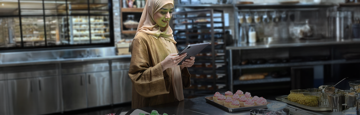 The image shows a woman in a kitchen wearing a hijab and holding a tablet, with trays of desserts in front of her.