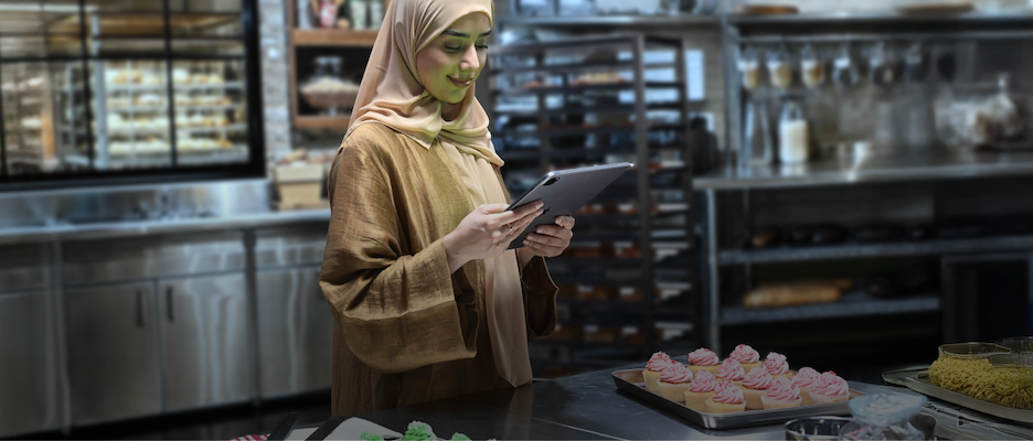 الصورة تُظهر امرأة في المطبخ ترتدي حجابًا وتمسك جهازًا لوحيًا بينما أمامها صواني حلوى.